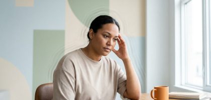 Femme assise à un bureau, main sur la tempe, l'air pensive ou souffrant de maux de tête. Une tasse et des cahiers sont visibles.
