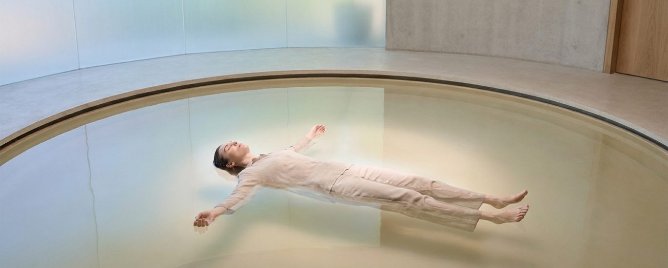 Une personne vêtue de vêtements clairs flotte paisiblement sur le dos au centre d'une piscine circulaire minimaliste.