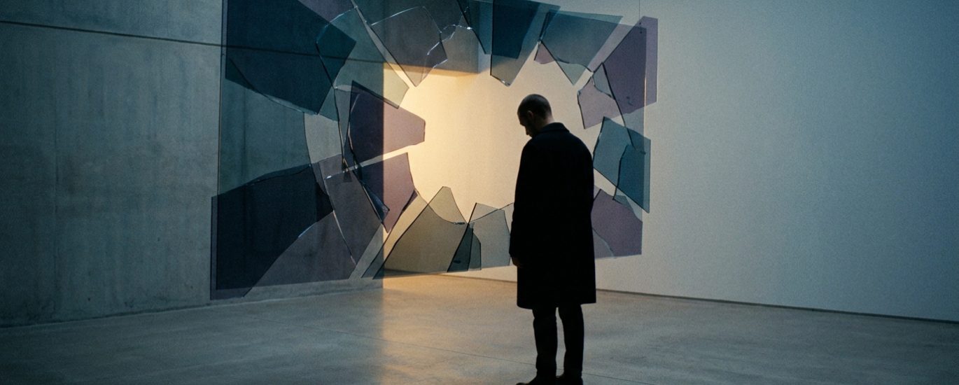 Un homme vêtu d'un manteau sombre observe une installation artistique composée de fragments de verre suspendus en l'air.
