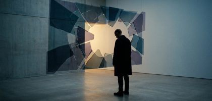 Un homme vêtu d'un manteau sombre observe une installation artistique composée de fragments de verre suspendus en l'air.