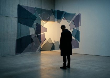 Un homme vêtu d'un manteau sombre observe une installation artistique composée de fragments de verre suspendus en l'air.